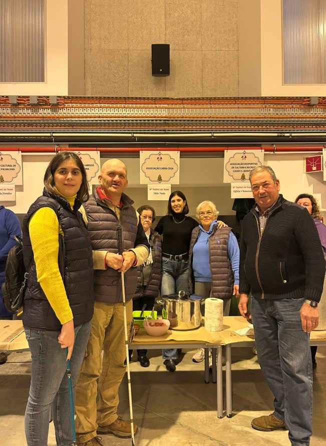 Grupo de pessoas em ambiente de convívio social, possivelmente numa feira ou encontro comunitário. Seis pessoas estão reunidas, todas estão a sorrir ou a mostrar uma expressão amigável. Algumas pessoas usam coletes acolchoados, e duas delas à frente seguram bengalas de orientação, sugerindo que são pessoas cegas ou com baixa visão. À frente do grupo há uma mesa com utensílios de cozinha, como uma panela de inox, taças verdes, uma taça com comida, rolo de papel e outros pequenos itens. No fundo, há placas penduradas com nomes de associações, como 